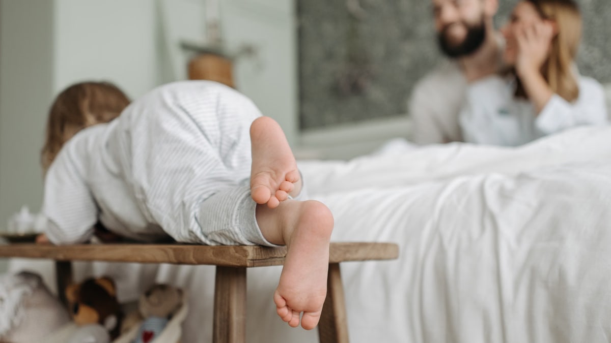Adorable child climbing in a cozy bedroom with parents in the background.